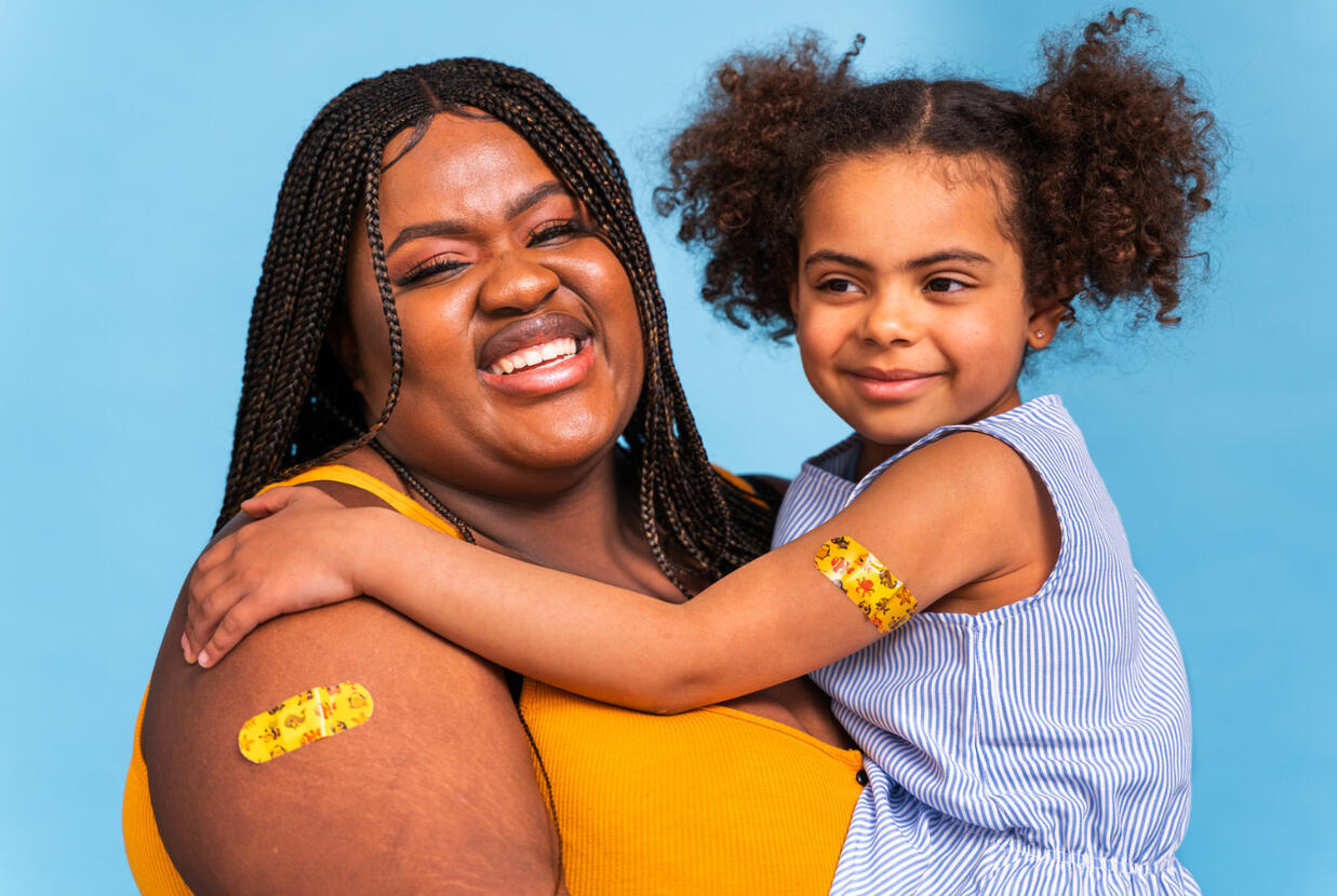 A smiling woman with teen who has a vaccine bandage on her shoulder sitting on her lap