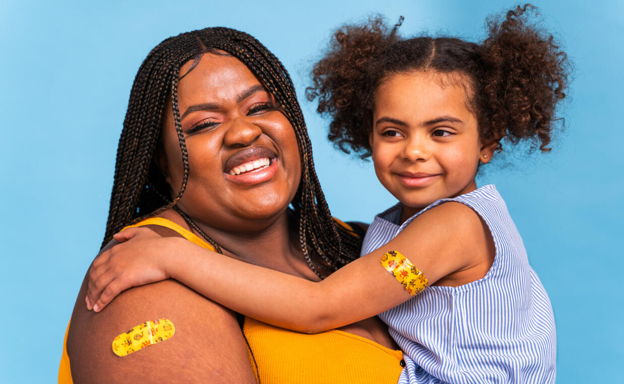 A smiling woman with teen who has a vaccine bandage on her shoulder sitting on her lap